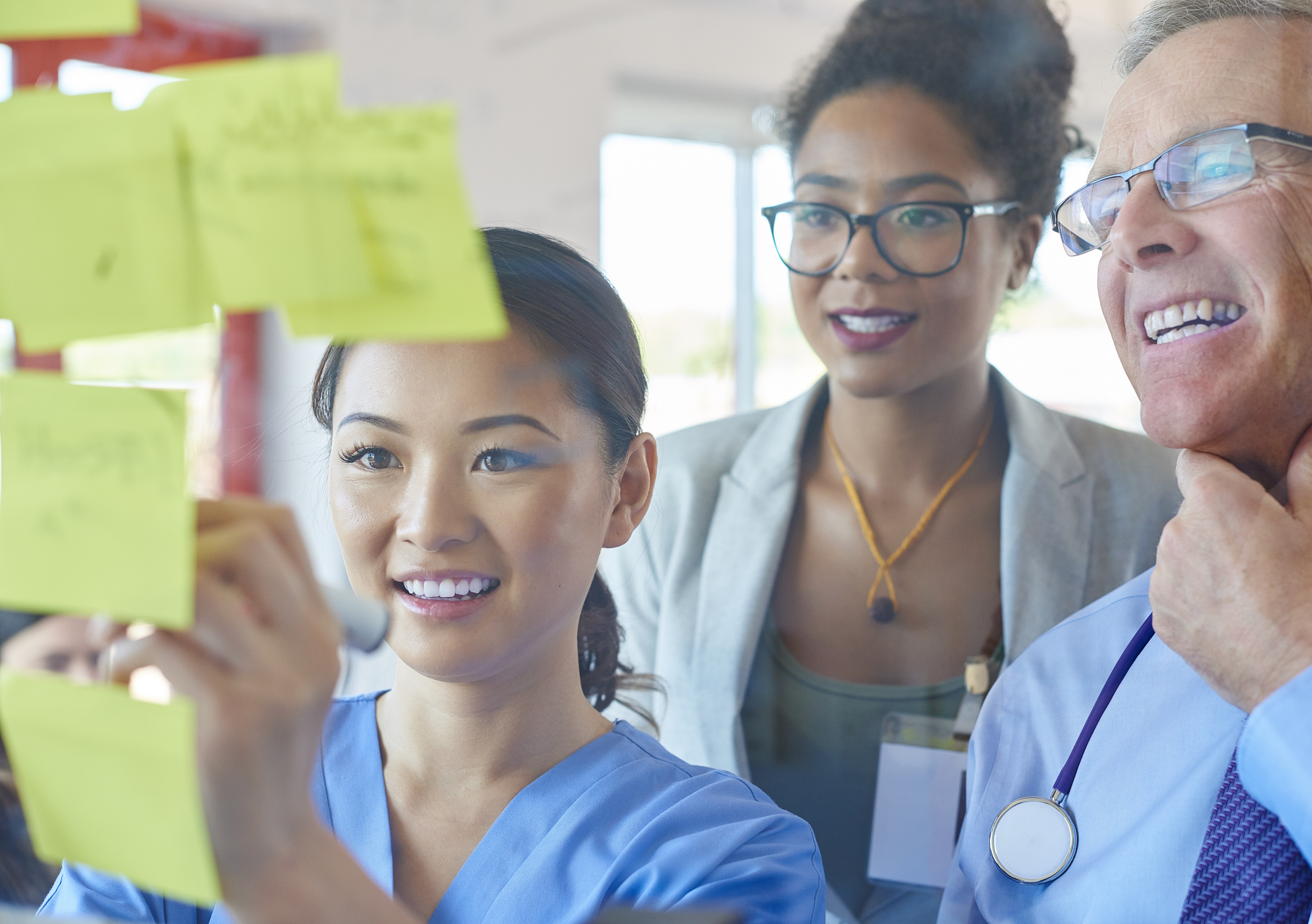 a senior doctor is looking at some ideas with a nurse . They are using stick notes on a glass display board in a medical meeting environment.